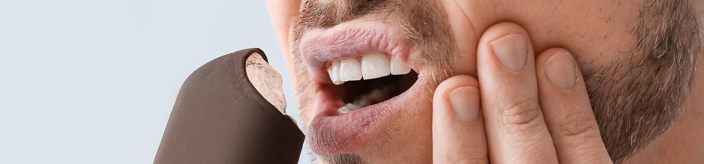 Homem com mão no rosto e expressão de dor ao morder um picolé, sugerindo sensibilidade em um dente só ao comer algo gelado.