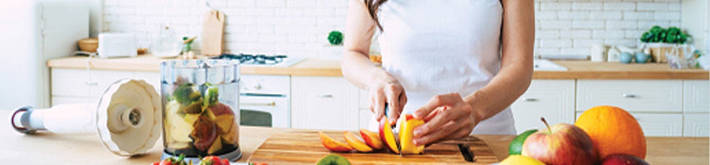 Mulher sorridente prepara uma refeição com frutas frescas em uma cozinha bem iluminada, promovendo uma alimentação saudável.