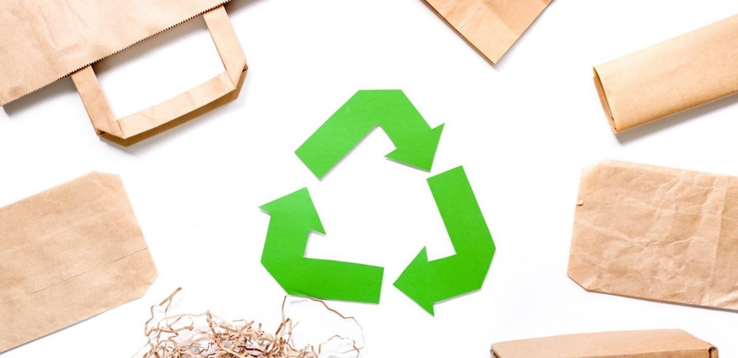 Símbolo verde de reciclagem cercado por embalagens e sacolas de papel kraft sobre fundo branco.