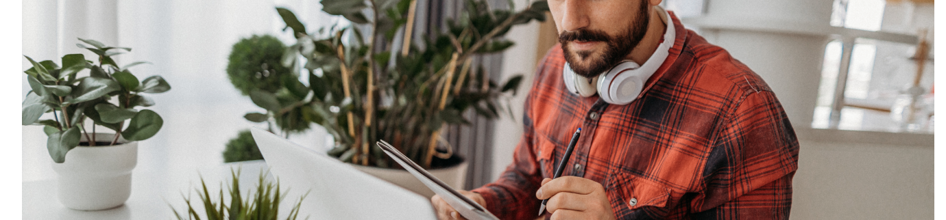 Homem em ambiente de trabalho com notebook e fones no pescoço, cercado por plantas, demonstrando concentração em uma tarefa.