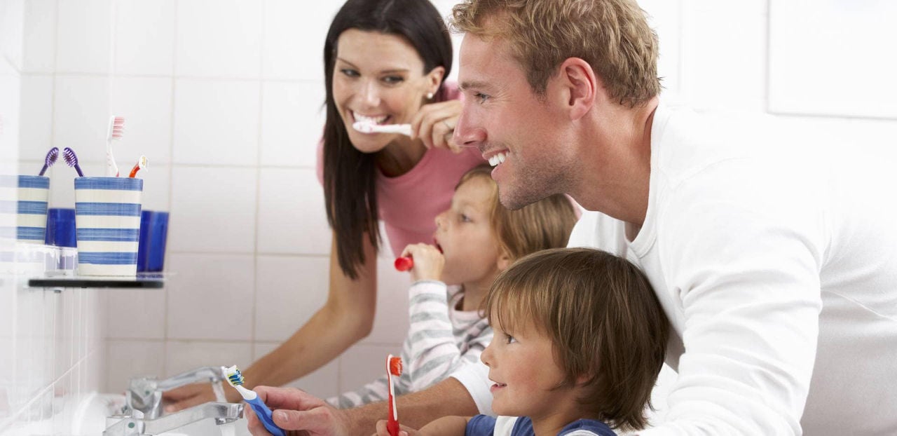 Família sorridente com homem, mulher e duas crianças escovando os dentes juntos no espelho do banheiro.